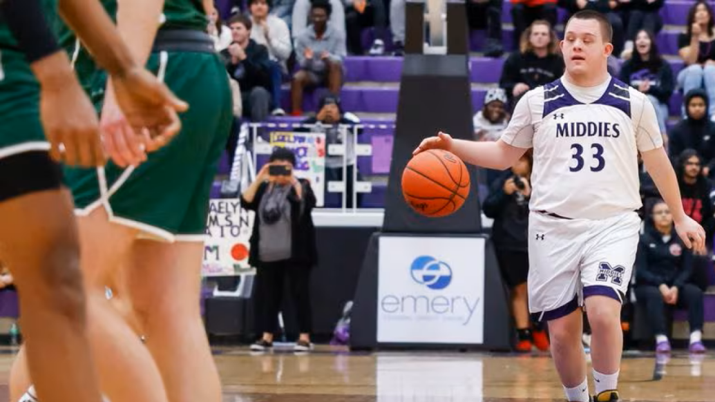 Boy with Down syndrome dribbles basketball down the court wearing a "Middies" jersey with the crowd cheering him on in the background.